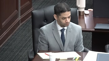 Un hombre vestido con traje gris y corbata azul sentado en una silla de oficina de cuero negro frente a un escritorio de madera oscura con papeles y lápices