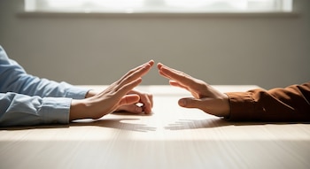 Dos manos, una con camisa azul y otra con camisa marrón, se alzan sobre una mesa de madera clara con luz natural. Están cerca pero sin tocarse.