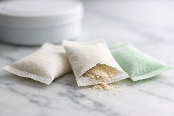 Tres bolsitas rectangulares, dos blancas y una verde claro, sobre una superficie de mármol. Una bolsita blanca está abierta con polvo beige claro visible.