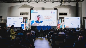Un auditorio con personas sentadas frente a un escenario iluminado, con pantallas grandes mostrando una oradora y logos, flanqueado por banderas de Guatemala y EE. UU