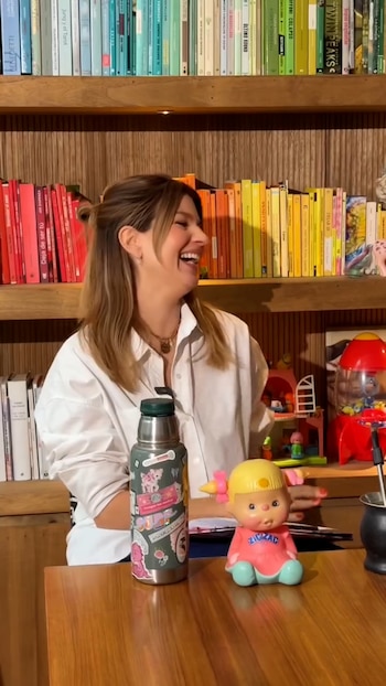 Una mujer sonríe sentada ante una mesa de madera con un termo decorado y un muñeco, delante de una estantería repleta de libros