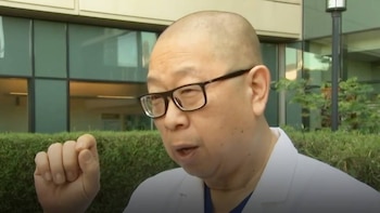 Hombre asiático calvo con gafas, vistiendo bata blanca, gesticula con su mano derecha levantada. Al fondo, un edificio de cristal y vegetación