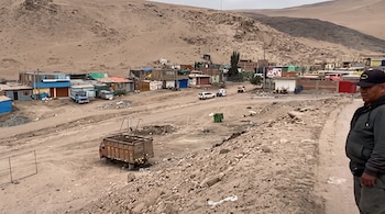 Un hombre sobre una pendiente rocosa junto a un camión, con casas y vehículos al pie de cerros de arena.