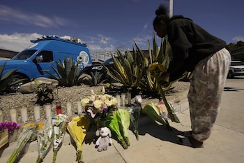 Una mujer coloca flores junto a ramos, velas y un peluche en la acera, ante plantas de agave y una camioneta azul de noticias