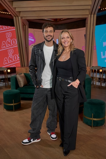 Diego Leuco y Carina Zampini posan de pie, sonriendo a cámara en un estudio de televisión con fondo de pantallas digitales y muebles verdes