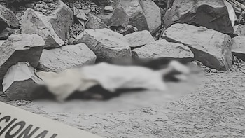 Cuerpo humano borroso tendido en el suelo de tierra con rocas alrededor, en una imagen monocromática. Se ve parcialmente una cinta de "POLICÍA NACIONAL"