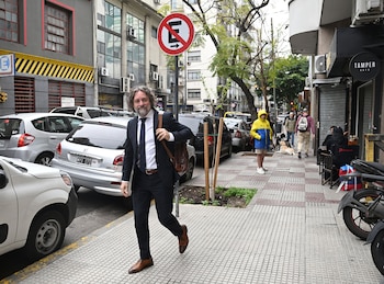 Un hombre con traje, corbata y bolso al hombro camina por una acera urbana junto a varios vehículos estacionados