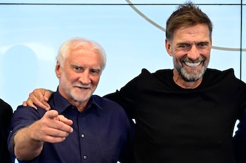 Dos hombres, uno con cabello y barba blancos, camisa azul, señalando con la mano derecha. El otro con cabello y barba castaños, camisa negra, sonríe. Fondo azul claro