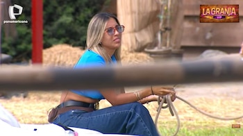 Gabriela Herrera, con cabello rubio y gafas, luce una blusa azul corta y jeans, sentada con expresión molesta en un set al aire libre con fardos de heno