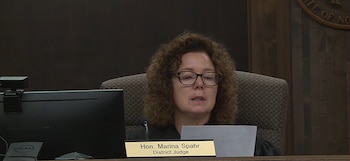 Primer plano de la jueza Marina Spahr con cabello rizado y gafas, sentada detrás de un monitor, con un cartel de identificación en el estrado
