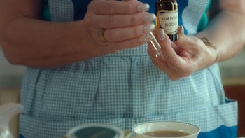 Manos con manicura clara sostienen un frasco marrón con etiqueta "CIANURO SODIO" y un gotero con una gota. Visten un delantal a cuadros azules