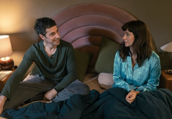 Esteban Lamothe y Griselda Siciliani sentados en una cama. Él sonríe a la izquierda, ella mira al frente a la derecha, ambos cubiertos por una manta oscura