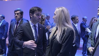Karina Milei conversa de perfil con un hombre con traje oscuro en un salón con iluminación azul rodeados de otras personas