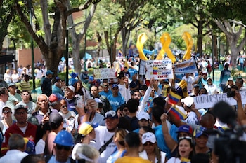 Multitud de personas marchando en una calle arbolada con carteles, banderas de Venezuela, algunas gorras y lazos amarillos grandes de fondo