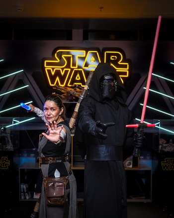 Una mujer vestida como Rey con sable azul y un hombre como Kylo Ren con sable rojo posan frente a un letrero de neón de Star Wars
