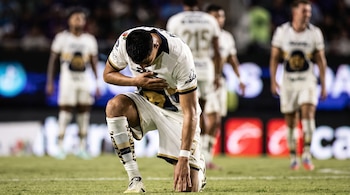 Futbolista hincado en la cancha con la mano en el pecho y la cabeza inclinada, mientras otros jugadores permanecen de pie en el fondo.