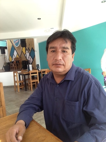 Primer plano de Rhudy Benavides sentado en una mesa de madera, vistiendo una camisa azul oscuro. Al fondo, un escenario con decoración geométrica y altavoces