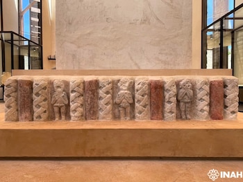 Relieve de piedra esculpida con tres figuras humanas de pie e inscripciones verticales entrelazadas y pintadas sobre un pedestal