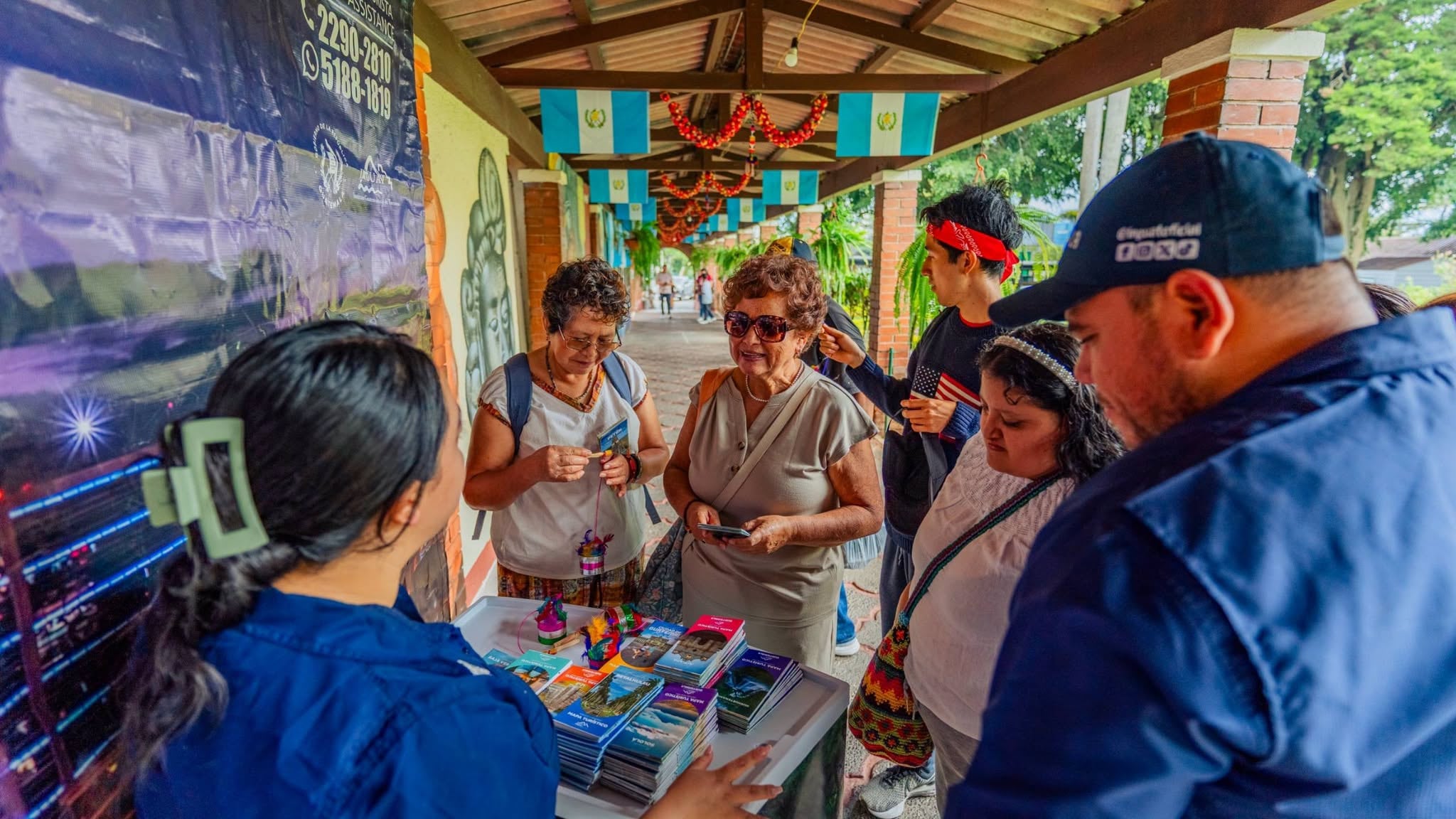 Un grupo de personas consulta folletos y mapas sobre destinos en un puesto de información turística decorado con banderas de Guatemala. (INGUAT)