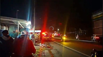 Calle nocturna con un camión de emergencia con luces encendidas, personas en la acera y motocicletas en circulación