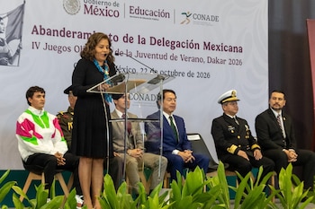 Una mujer habla ante un atril transparente acompañada por un atleta, mandos militares y funcionarios sentados frente a un panel oficial