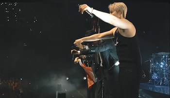 Tres músicos en un escenario oscuro. Uno, rubio y de camiseta negra, alza un micrófono. Otros dos sostienen micrófonos. Se ve una tela roja