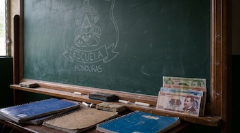 Un pizarrón verde con marco de madera, dibujo de escudo y texto "Escuela Honduras", tizas, borradores, cuadernos y billetes de lempira apilados.