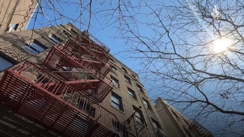 Edificio de apartamentos de ladrillo con escaleras de incendio rojas en su fachada, visto desde abajo. Ramas de árboles desnudas y un cielo azul con sol brillante