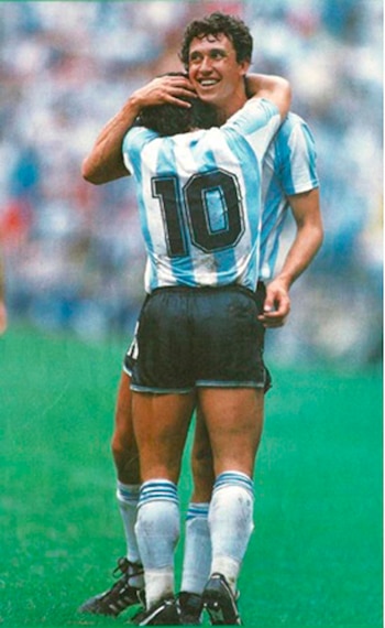 Dos hombres vestidos con uniformes de fútbol de Argentina se abrazan en un campo de césped. Uno lleva el número 10 en la espalda. Llevan pantaloneta negra