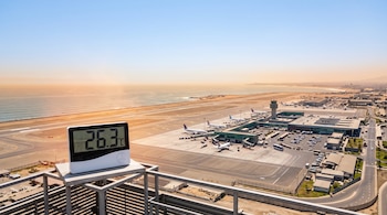 Un termómetro digital marca 26,3 °C sobre una estructura metálica frente a la pista de aterrizaje y terminal de un aeropuerto.