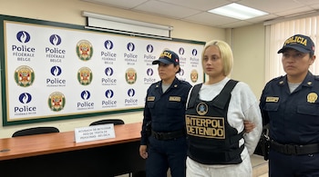 Mujer con chaleco "INTERPOL DETENIDO" es escoltada por dos policías uniformadas frente a un panel con logos de la Policía Federal Belga y un cartel.