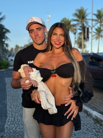 Un hombre con gorra sostiene a un bebé junto a una mujer vestida de negro en una acera al aire libre con palmeras al fondo