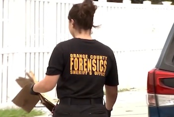 Espalda de una persona con camiseta negra de "Orange County Forensics Sheriff's Office", guantes negros, un objeto marrón en la mano, valla blanca y parte trasera de un coche