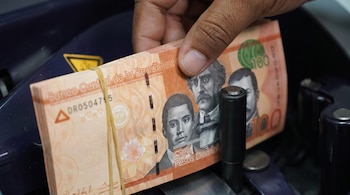 Una mano sostiene un fajo de billetes anaranjados de cien pesos dominicanos sujeto con una goma elástica dentro de una máquina contadora.