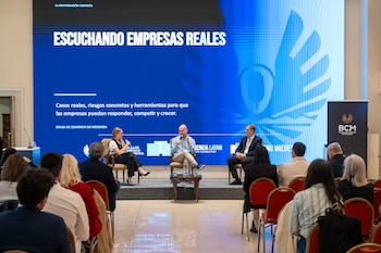Tres personas sentadas en un escenario hablan frente a un público, ante una gran pantalla azul con texto y un panel lateral