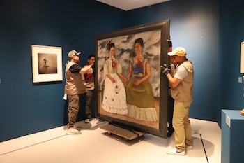 Cuatro personas con chalecos y guantes mueven una pintura de "Las dos Fridas" con un marco oscuro sobre un patín en una sala de galería de paredes azules. Otro cuadro cuelga en la pared