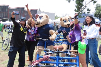 Varias personas, una con máscara de Batman y dos con disfraces de puma, posan para fotos en un evento de ciclismo al aire libre junto a bicicletas tándem azules