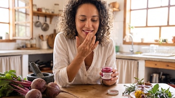 Mujer con cabello rizado aplica bálsamo rojo en sus labios con el dedo. Sostiene un frasco de "Bálsamo de Betabel Natural". Hay betabeles en la mesa.