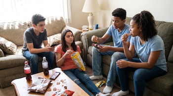 Cuatro adolescentes sentados en sofás y suelo consumiendo patatas fritas, galletas saladas, chocolate y bebidas.