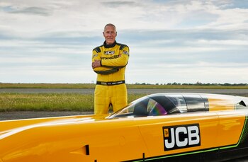 Andy Green, con traje de carreras amarillo, de pie junto a un vehículo de velocidad terrestre JCB Hydromax amarillo y negro, en una pista y campo abierto