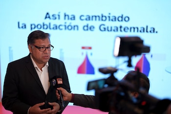 Hombre en traje y gafas habla a micrófono. Pantalla de fondo con texto 'Así ha cambiado la población de Guatemala' y pirámide demográfica. Cámara y foco de luz