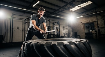 Un hombre musculoso con camiseta gris oscuro golpea un neumático negro grande con un mazo pesado en un gimnasio. Se observan luces brillantes y un fondo de ladrillo.