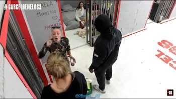 Vista aérea de una participante de Gran Hermano gesticulando enérgicamente; detrás de barrotes, otra mujer está sentada en una cama en una celda