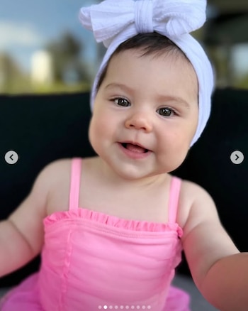 Primer plano de un bebé de tez clara, ojos oscuros y cabello oscuro, sonriendo ampliamente, llevando una diadema blanca y un traje de baño rosa