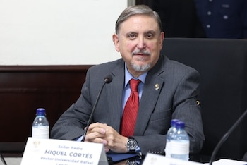Hombre con traje gris y corbata roja sentado a una mesa. Hay un micrófono, botellas de agua y una placa de identificación con el nombre Miquel Cortés
