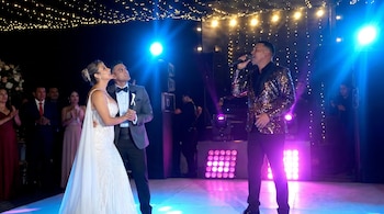 Cantante con chaqueta de lentejuelas en escenario, frente a pareja vestida de boda en pista de baile iluminada con luces azules y guirnaldas brillantes.