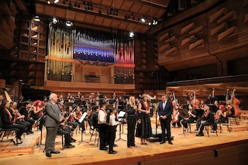 Músicos de una orquesta sentados en un escenario de madera frente a un gran órgano, con varias personas de pie al frente