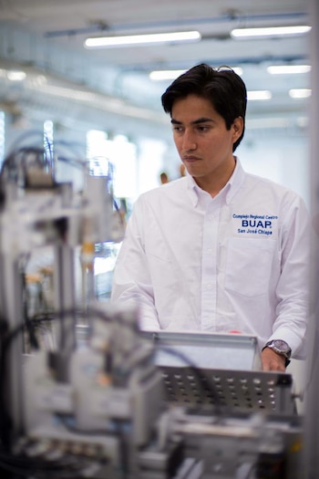 Un hombre joven de cabello oscuro y camisa blanca con el logo de "BUAP San José Chiapa" mira hacia abajo a equipos tecnológicos en un laboratorio iluminado