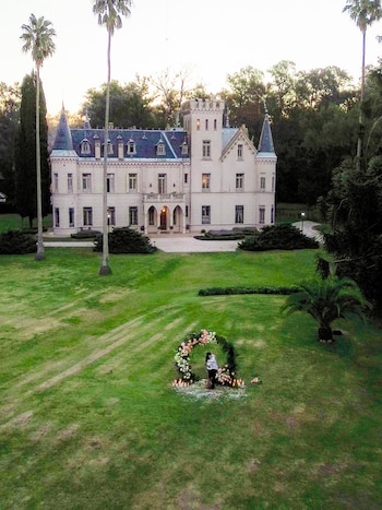 Vista aérea de una pareja abrazándose bajo un arco floral con velas, en un gran jardín verde frente a un castillo señorial al atardecer