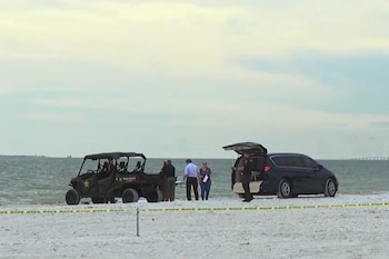 Playa de arena con dos vehículos policiales, varias personas, cinta amarilla de acordonamiento y el mar de fondo bajo un cielo nublado